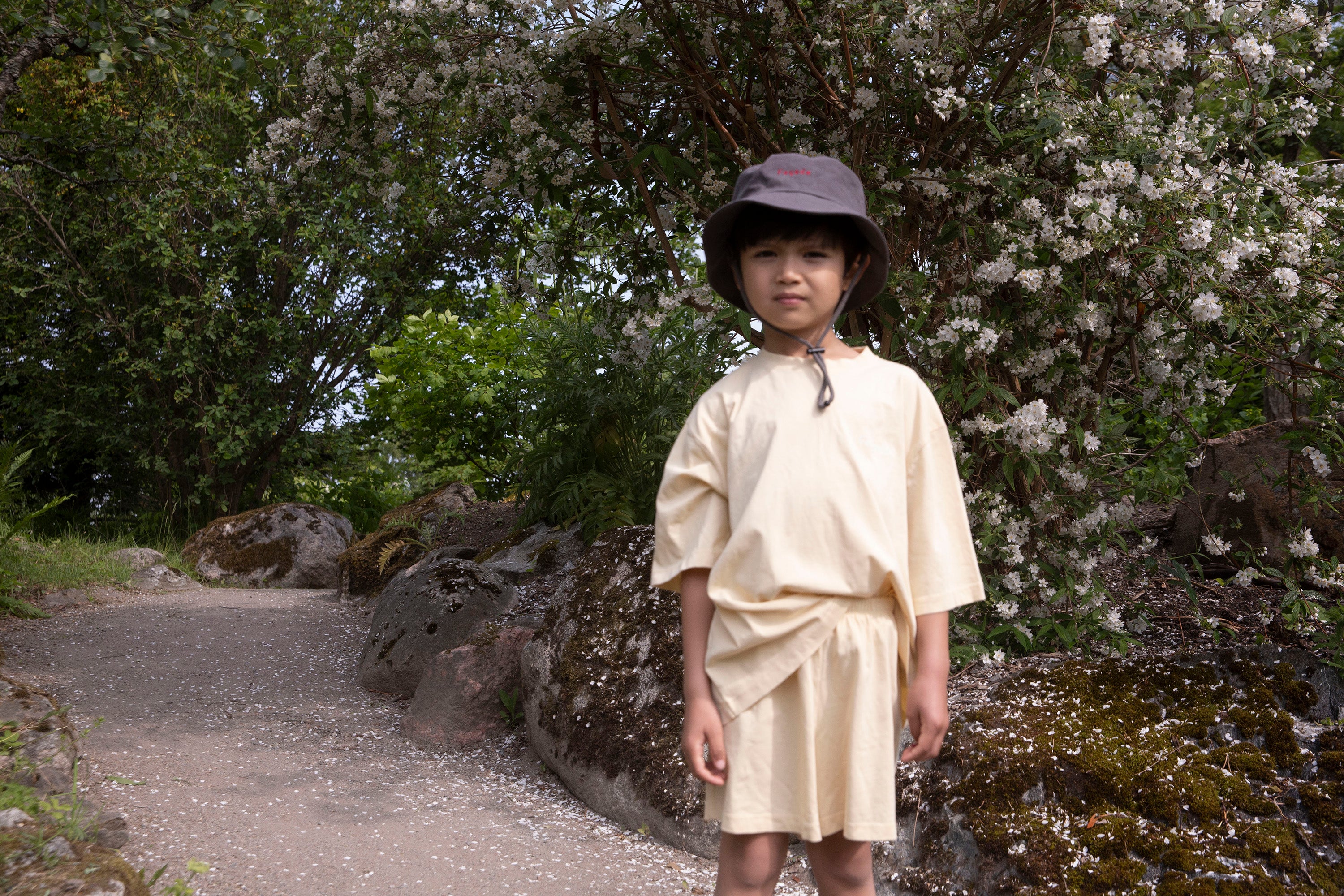 Child wearing Façade Bato Wide Tee, Mido Runner Shorts and Refu Camping Hat in butter and asphalt, organic cotton kids outfit photographed outdoors