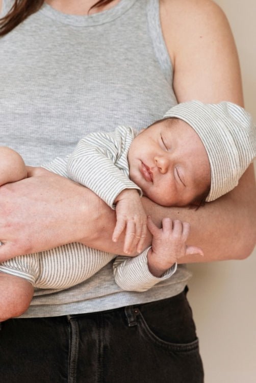 Baby sleeping in mothers arms. Baby is wearing a gray label bodysuit and a beanie