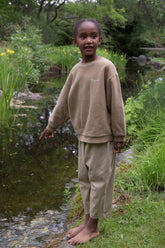 Child wearing Façade Kohi Wide Pants in hay, organic cotton kids trousers with a relaxed barrel fit, standing outdoors near a pond