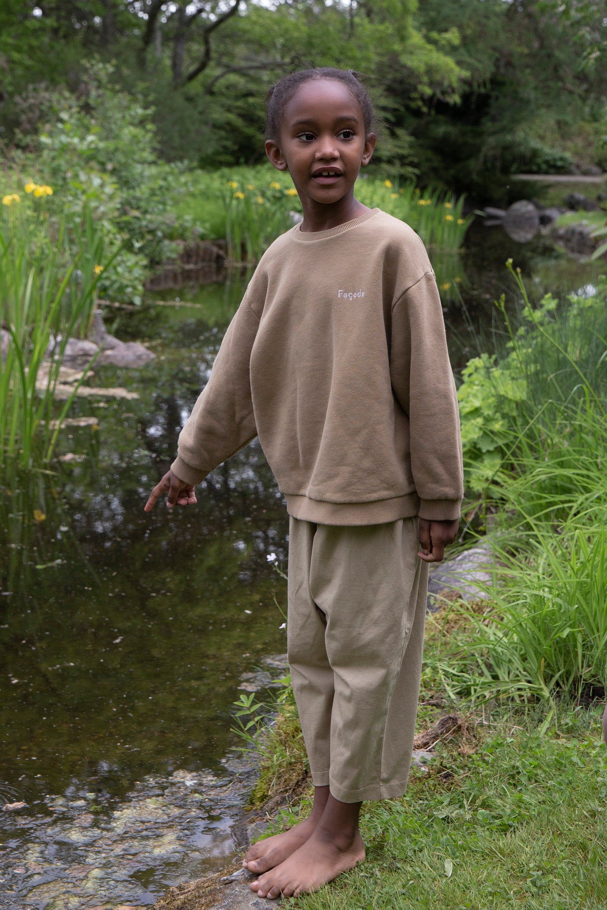 Child wearing Façade Kohi Wide Pants in hay, organic cotton kids trousers with a relaxed barrel fit, standing outdoors near a pond