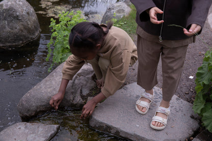 Child wearing Façade Kohi Wide Pants in mole brown, organic cotton kids trousers with a relaxed barrel fit, worn outdoors by the water